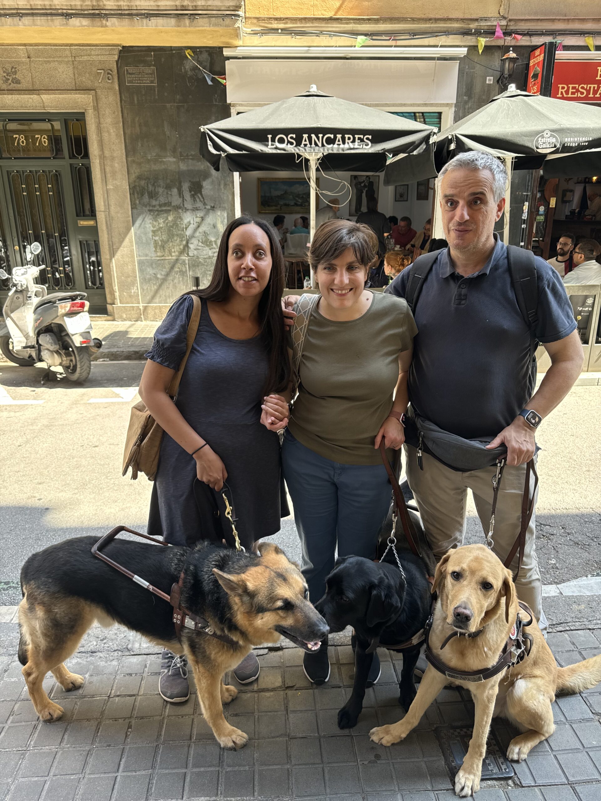 Video: Haben, Daniela, and her husband Jos&eacute; stand together on a sunny sidewalk. Haben wears a blue dress, Daniela wears a green shirt and jeans, and Jos&eacute; wears a Navy blue shirt, khakis, and an Apple Watch. Next to German Shepherd Mylo is Rosie, a black lab, and beside Jos&eacute; sits a yellow lab sweetly looking at the camera. Behind the smiling group, across the street, is a warm restaurant with indoor and outdoor seating.