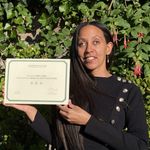 Haben enthusiastically holds up a certificate that says, “La Ministre de la Culture nomme par arrêté de ce jour. Madame Haben Girma: Chevalière de l’Ordre des Arts et des Lettres.” It is signed by Rachida Dati, Ministre de la Culture, and the signature has a tactile seal over it. Haben is wearing a long-sleeved black top with gold buttons along one side. Behind her is a wall of hanging fuchsias.