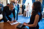 In the Blue Room at the White House celebration of the 25th Anniversary of the Americans with Disabilities Act, President Barack Obama is standing at a table typing to Haben Girma using a white QWERTY keyboard. He’s wearing a charcoal suit with a blue tie and a smartwatch. Haben is across the table from him reading his words in Braille on her BrailleNote Apex. She’s wearing a sleeveless navy blue dress. Vice President Joe Biden and Senior Advisor Valerie Jarrett observe the interaction with interest. In the background, the room has a vibrant blue rug with gold star patterns and matching floor-length drapes. Painted portraits adorn the walls.
