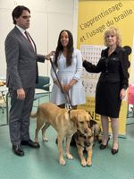 President Matthieu Juglar and his guide dog Rider, Haben Girma and her guide dog Mylo, and Director General Carole Godin. They are standing in front of a vertical yellow banner with black text, &ldquo;L'apprentissage et l'usage du braille.&rdquo; A chart shows the Braille and print alphabets and numbers. Beside that, six large circles visually represent a standard Braille cell. The banner also shows Braille music notation.