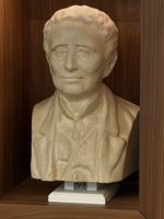 A polished wood display shelf has a pedestal holding a stone-like bust of Louis Braille. He wears a calm expression with his eyes closed, his wavy hair cropped short, and a collared shirt under a coat. The pedestal has a QR code on it, though part of it is not visible.