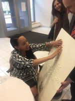 Damian Sian holds up a Princeton University jumbo braille poster as Haben reads it with two hands. She's sitting on a chair with a braille computer on her lap