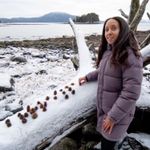 Brailling on a beach in Alaska. A small, secluded island full of evergreens sits in the middle of Gastineau Channel, and on the far shore rise mountains. I'm smiling as I hold a cone on top of a large, snow-covered log. The log has twenty-five Spruce and Hemlock cones half submerged in snow. The cones spell: brrraille! Photography by Cameron Byrnes.