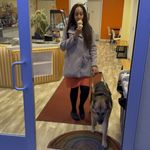 Haben and her Seeing Eye dog walk out of a cafe with a rainbow welcome mat by the door. She has one hand on Mylo's harness and the other holding a golden waffle cone topped with ice cream.