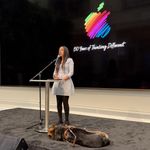 Haben Girma, a woman with dancing hazel eyes, long black hair, medium dark skin, and wearing a blue and white dress, stands on stage speaking into a microphone. A round table in front of her holds a Braille display where her fingers gently read. Next to her, stretched out on the floor, rests a German Shepherd Seeing Eye dog. Behind them is a black screen with a rainbow Apple logo above the words, “50 Years of Thinking Different.”