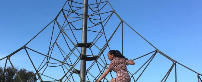 Haben climbs a large pyramid-shaped jungle gym that has multiple routes to the top. She is about two thirds of the way up the 20-foot high rope structure. Trees and blue skies are in the background.