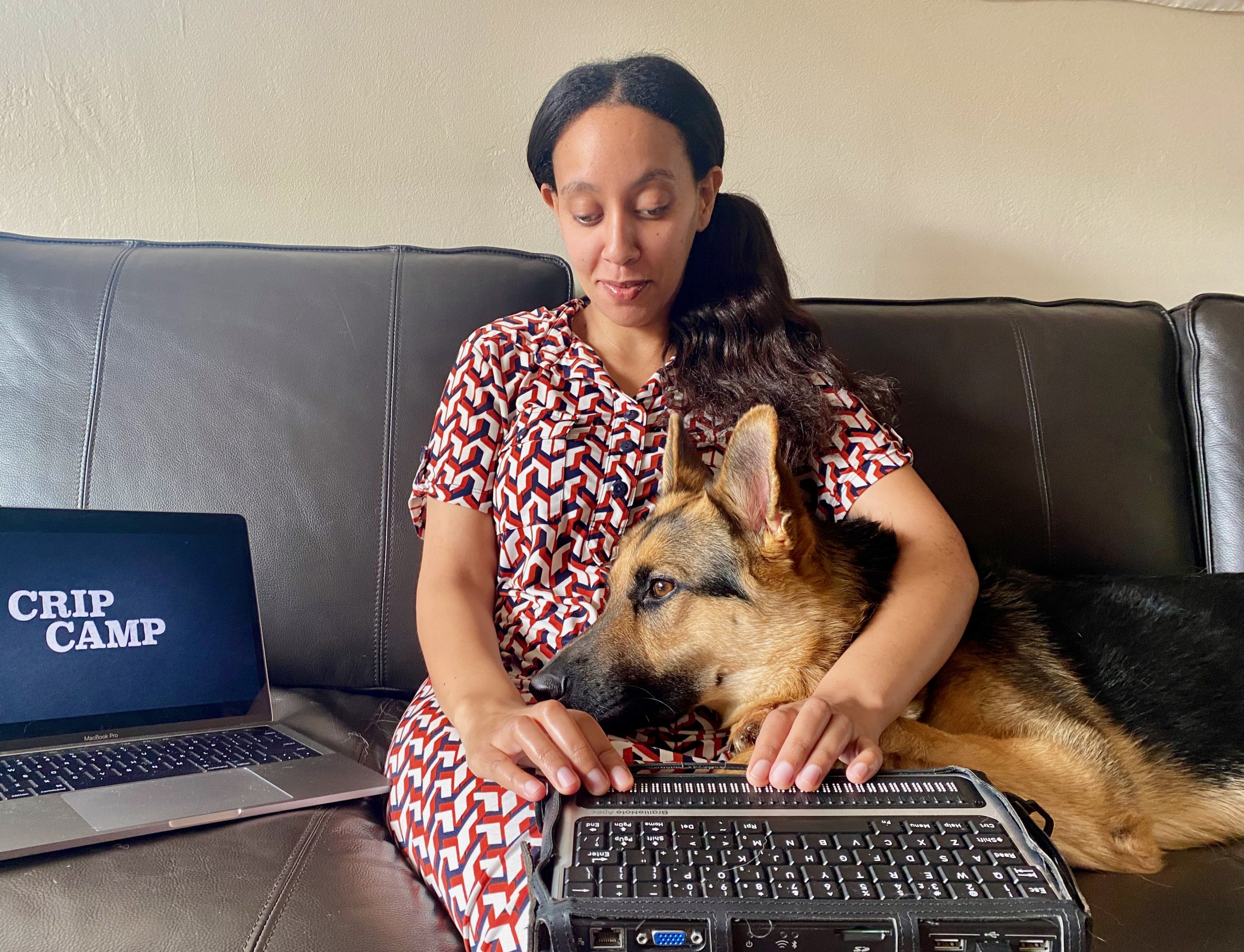 Haben is sitting on a sofa beside a laptop showing Crip Camp. Her braille computer is on her lap, as well as the head of Mister Mylo the German Shepherd who is attentively watching the screen