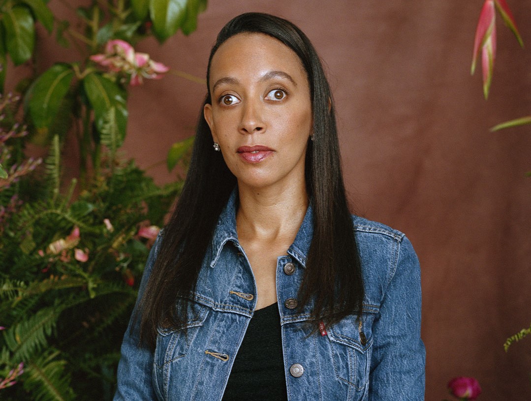 I, Haben, a Black woman with long black hair, am sitting on a stool in front of a brown background and green leaves with pink floral arrangements. I’m wearing a denim jacket over a black top and white patterned skirt