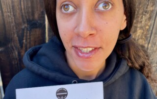 I’m wearing a black hoodie with the book club’s name Underrated on the left side. I’m smiling and holding up the card. A dark brown fence is in the background.