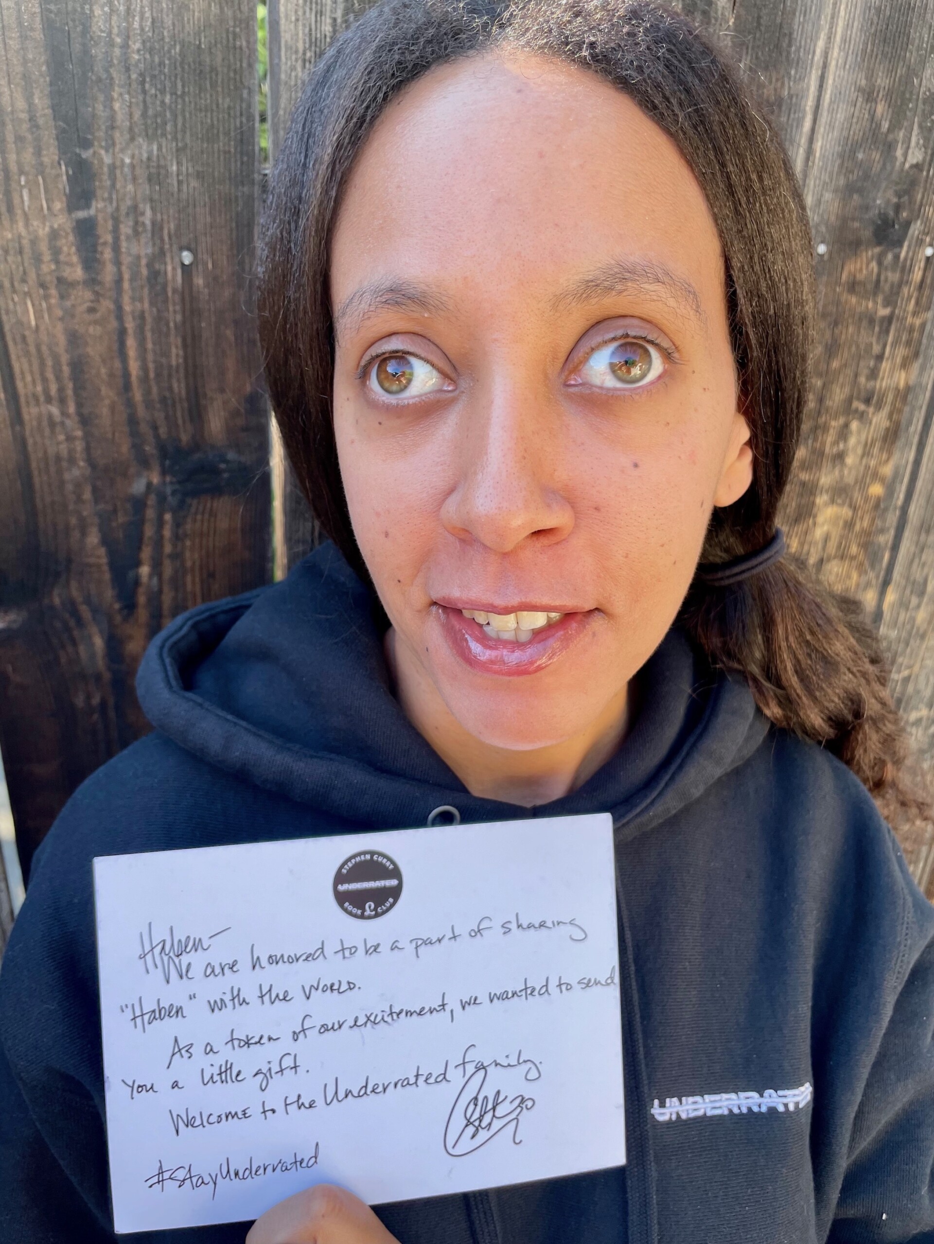 I’m wearing a black hoodie with the book club’s name Underrated on the left side. I’m smiling and holding up the card. A dark brown fence is in the background.