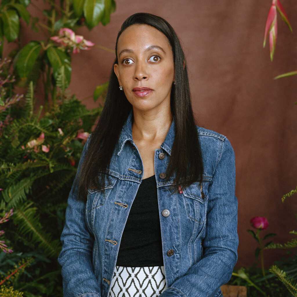 I, Haben, an Eritrean American woman with long black hair, am sitting on a stool in front of a brown background and green leaves with pink floral arrangements. I'm wearing a denim jacket over a black top and white patterned skirt.