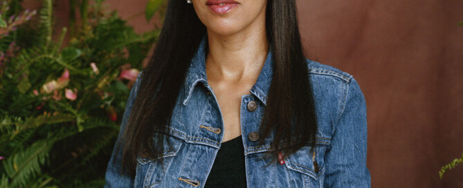 I, Haben, an Eritrean American woman with long black hair, am sitting on a stool in front of a brown background and green leaves with pink floral arrangements. I'm wearing a denim jacket over a black top and white patterned skirt.