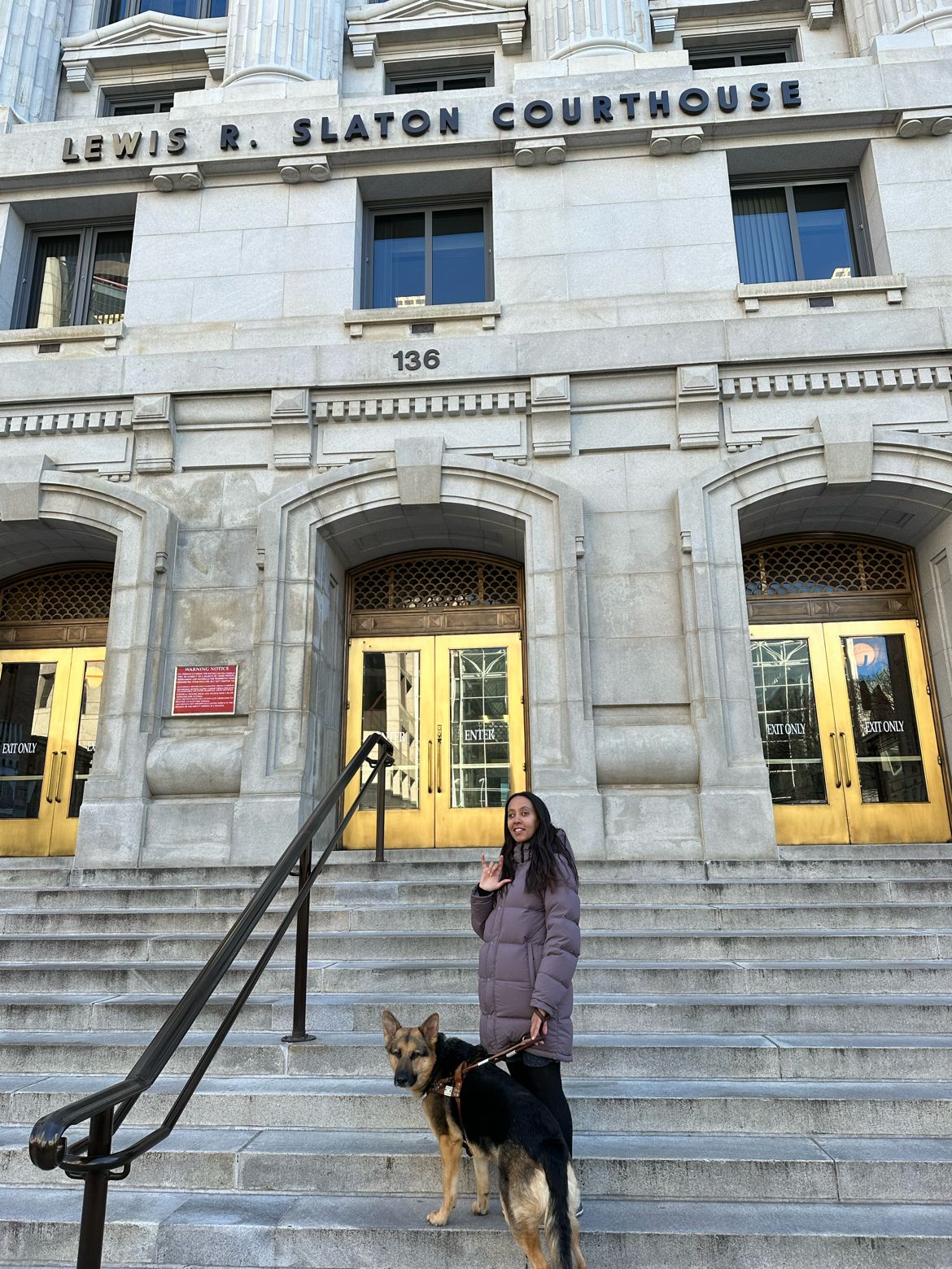 Seeing Eye dog Mylo and I stand on the steps of the Lewis R. Slaton Courthouse. I’m smiling and signing ILY. Behind us, the courthouse doors glow golden in the sun.