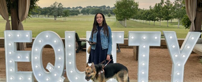 Haben and her guide dog Mylo standing in front of giant light-up letters spelling "Equity" with beautiful green fields in the background.