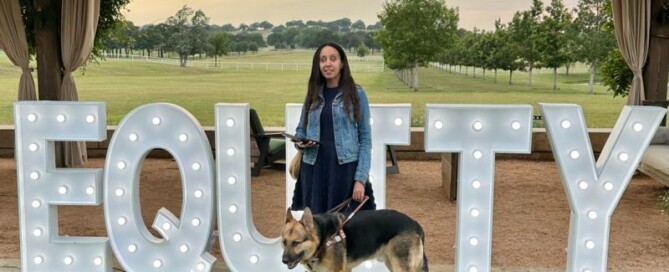My Seeing Eye dog and I stand in front of a large sign. About four feet tall, the 3D letters spell EQUITY. Lightbulbs on each letter brighten the word. Behind them stand a row of trees, beyond which stretches a lush green field topped by clouds glowing in the setting sun.