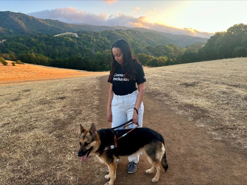 I’m wearing a shirt that says “Commit to Inclusion” and smiling down at Mylo, a German Shepherd Seeing Eye dog. Behind us is a grass-covered hillside with forested hills in the distance.