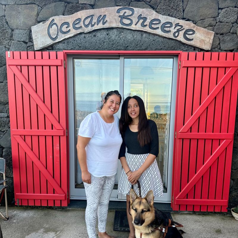Vibrant red shutters frame sliding glass doors, crowned by a curving sign reading, “Ocean Breeze.” Dalila and I stand smiling in front of the doors, and Mylo is sitting beside us.