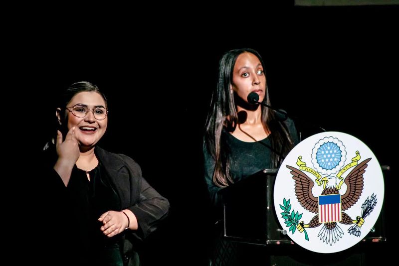 Close up of me at the podium that has a U.S. Seal. Beside me stands the LSM interpreter (Mexican Sign Language).