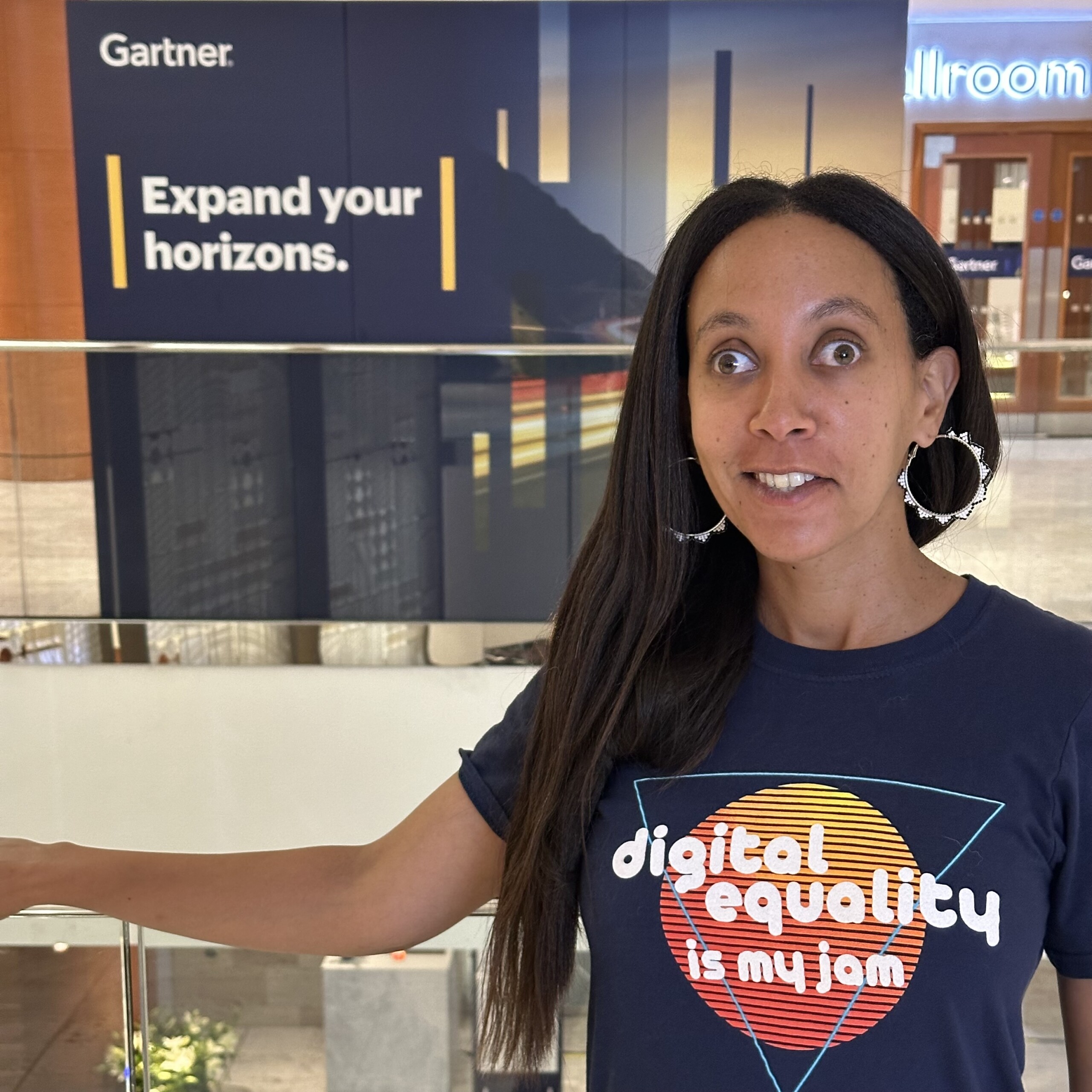 I’m smiling and wearing a shirt with text that says, “Digital equality is my jam.” Behind me, across the hotel lobby, a large Gartner sign with a stylized coastline reads, “Expand your horizons.”