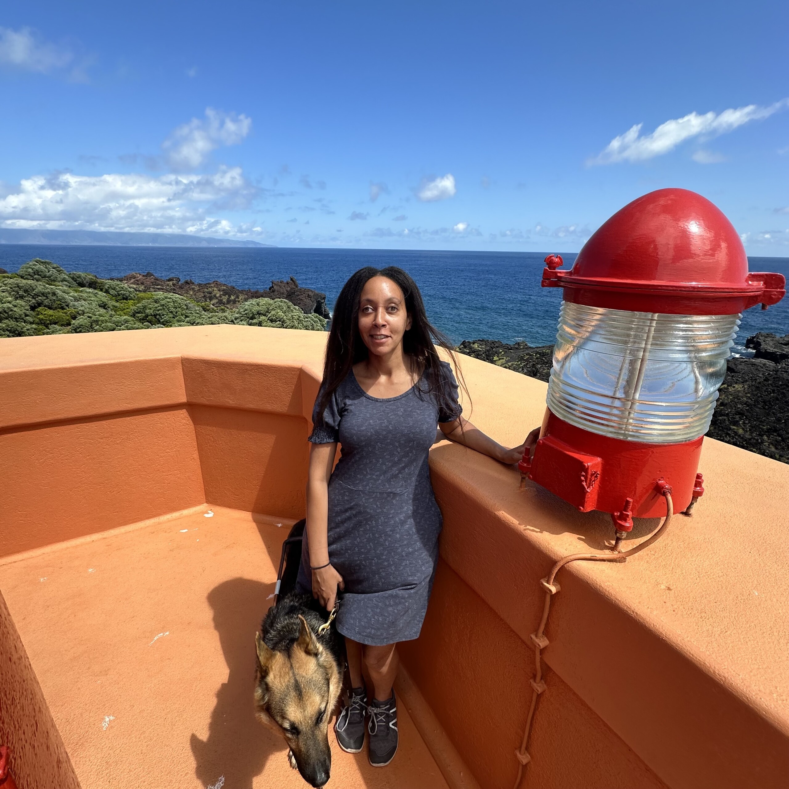 High up on the tower’s viewing platform, I stand beside an orange, waist-high wall where the reserve light happens to be perched. The large lantern has red along its top and bottom, and a silvery, translucent center. My guide dog Mylo looks alert beside me, and behind us, down below, appear the tops of trees, deep blue waters, and the shores of another island called São Jorge.