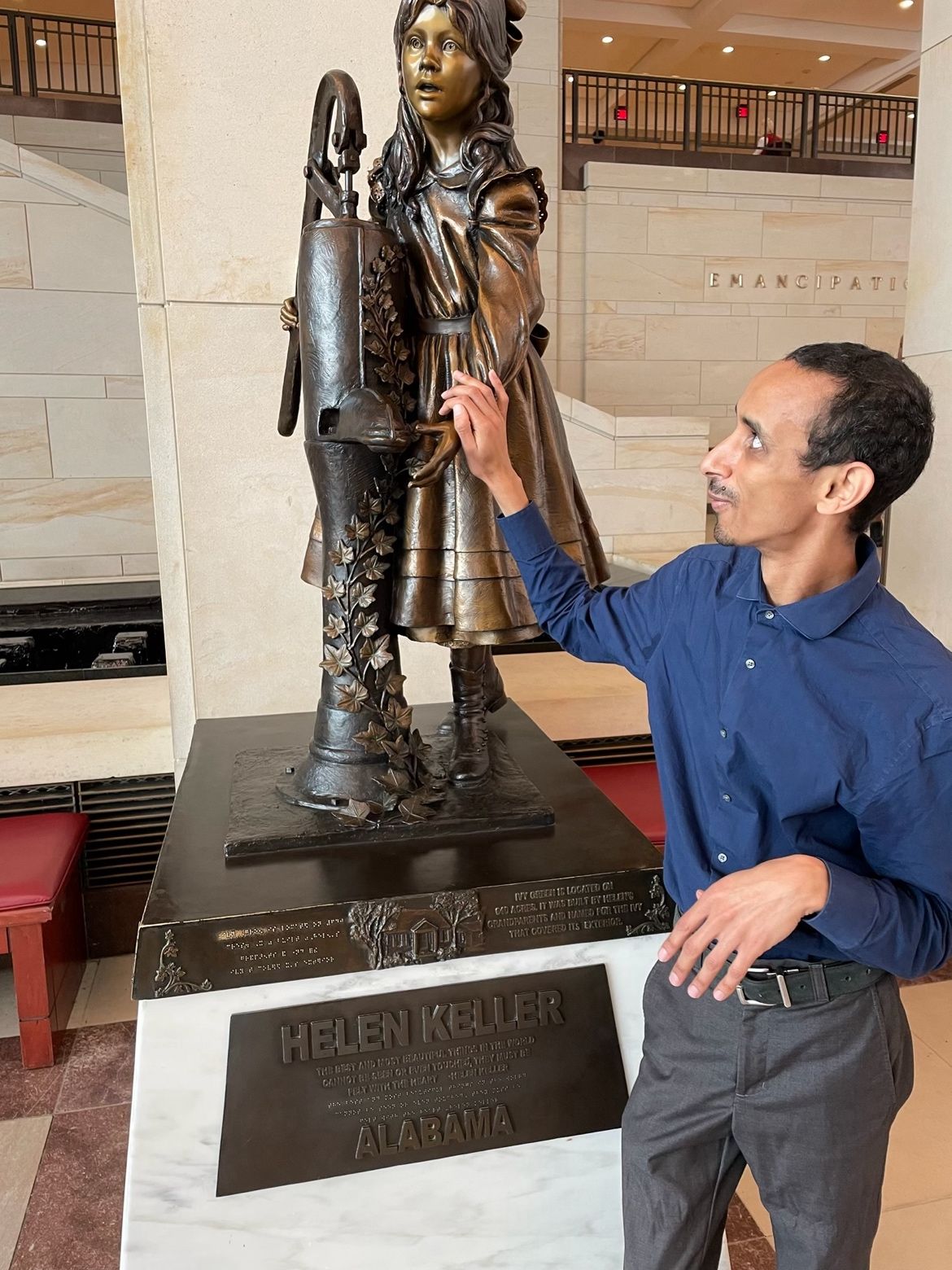 Mussie, a tall Eritrean American Deafblind man, stands beside the Helen Keller statue inside the U.S. Capitol. Young Helen has one hand on the handle of the water pump, and the other hand under the spout. Mussie studies Helen’s hand beneath the water spout.