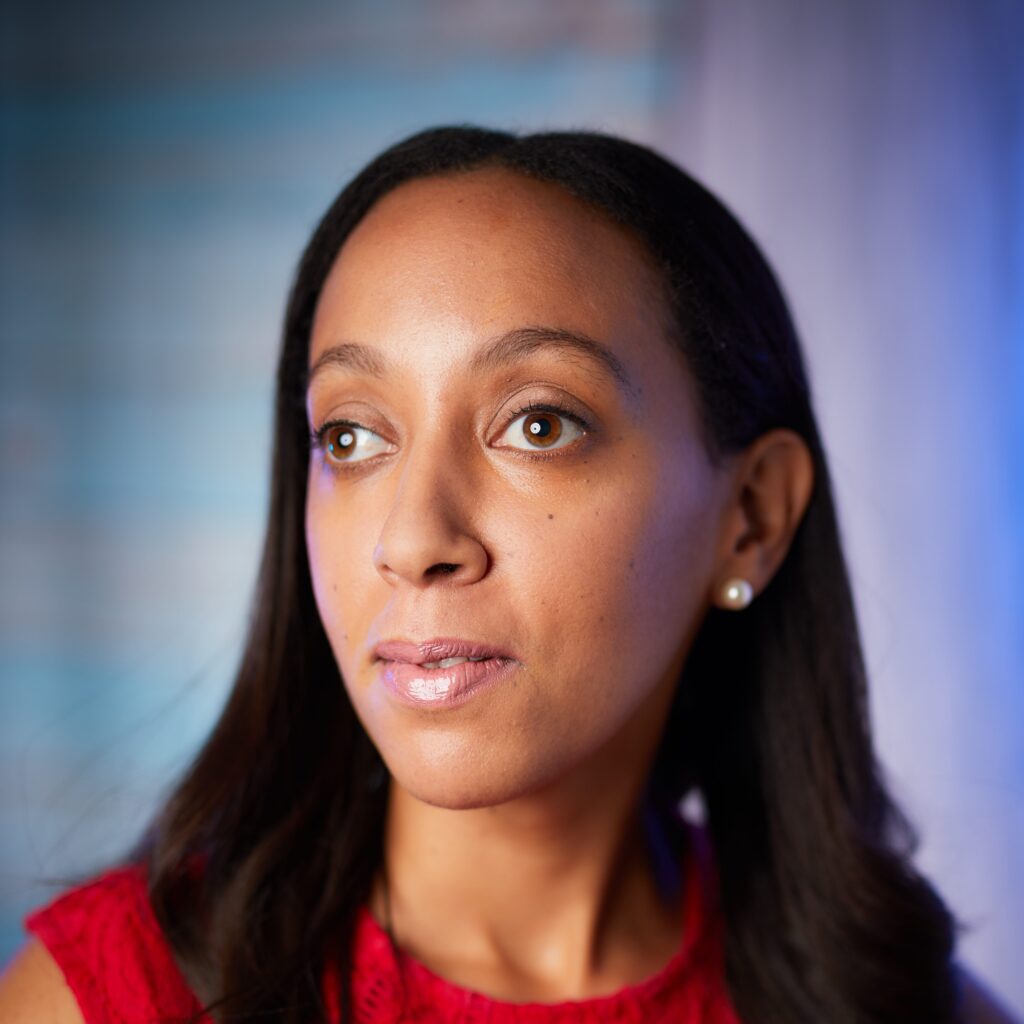 A portrait of Haben in front of a blue and purple backdrop. She is wearing a red dress, has pearl-shaped earrings, long black hair, and has a serious expression.