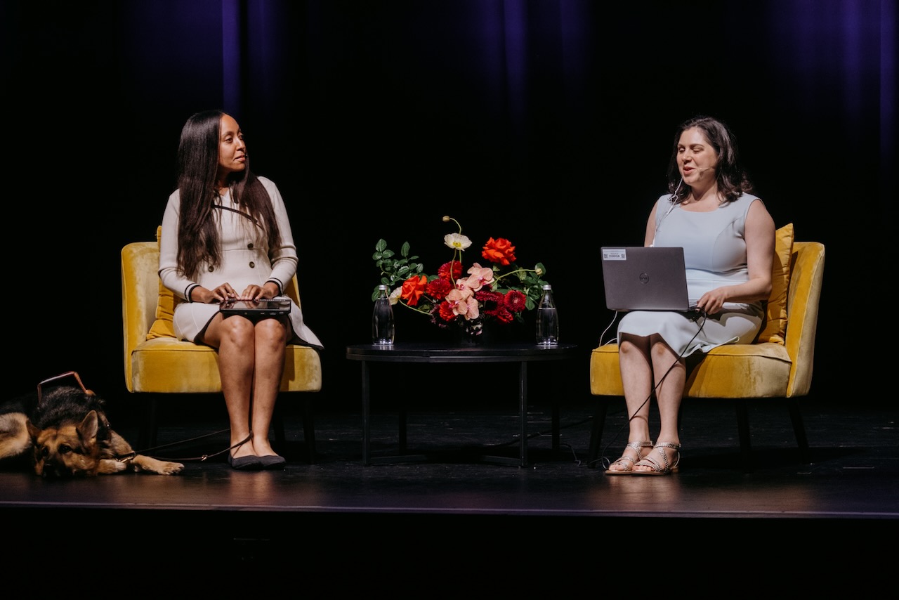 Two women sit on stage facing each other, and a small table with flowers is between their chairs. Nas has a laptop wired to an earpiece that allows her to read questions and notes using her screenreader. Haben sits with her fingers on a Braille computer, listening as Nas asks a question. Seeing Eye dog Mylo rests by Haben’s feet.