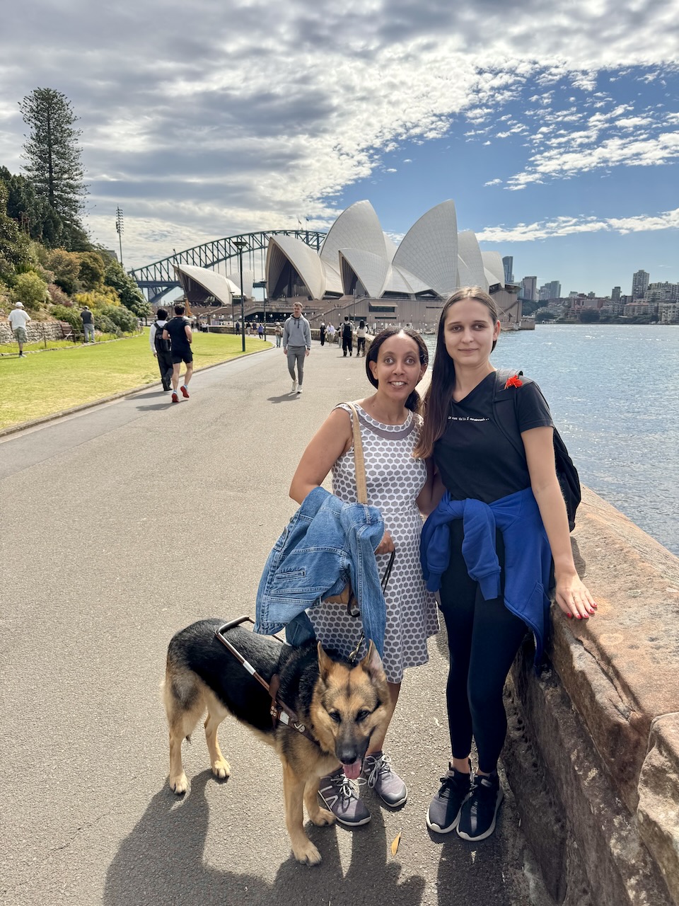 Haben and Seeing Eye dog Mylo stand next to a young woman. They’re all smiling, and behind them is the Sydney Opera House, the bright blue water of the harbor, and the bridge.