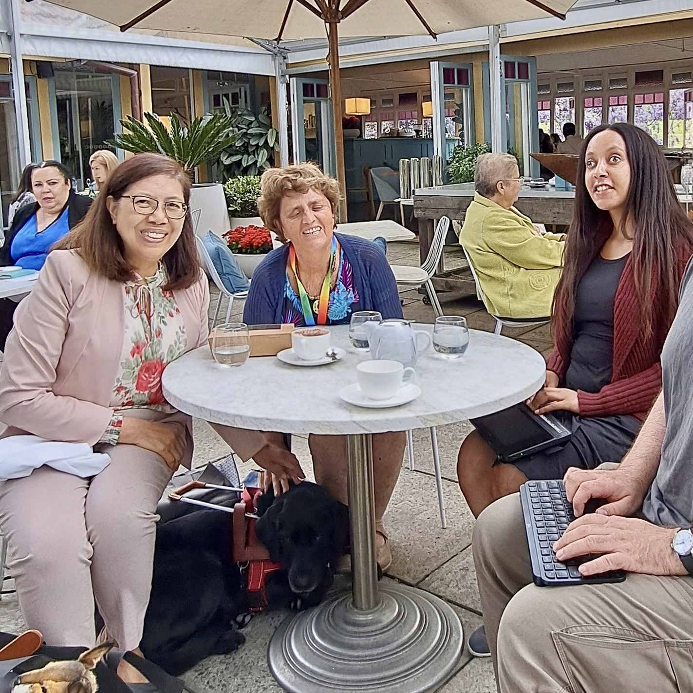 Three women sitting around a table in a plant-filled terrace, and they all seem to be enjoying coffee and drinks. On the left is Charissa, in the middle is Gisele, and her Labrador guide dog Nyota is under the table. On the right is Haben with a Braille display on her lap.