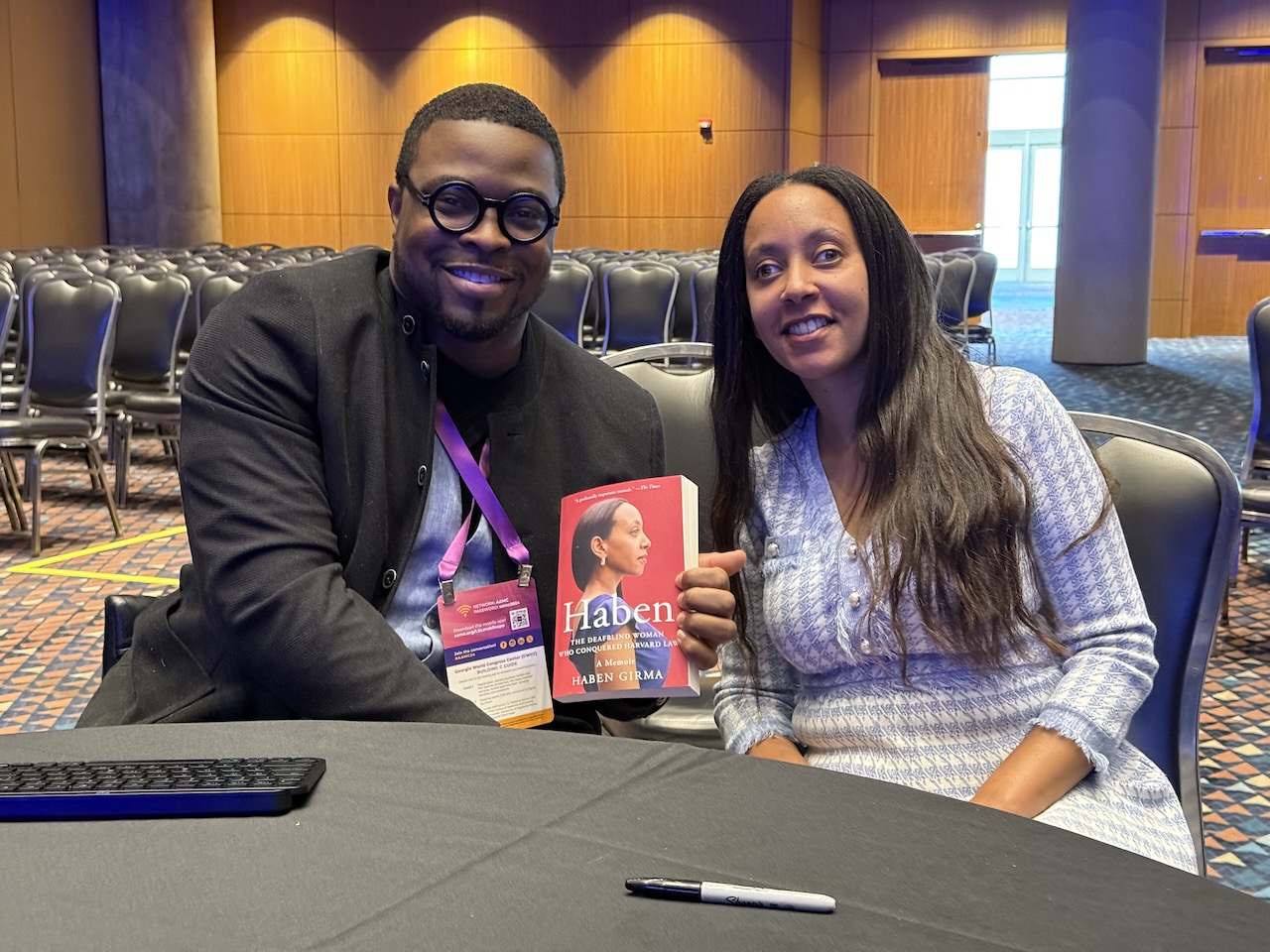 Dr. O smiles as he holds up a copy of Haben’s book, and she smiles as she leans toward him. Haben is a Black woman in her thirties wearing a blue dress and long, dark hair. Dr. O is a Black man in his thirties wearing a dark blazer, dark-rimmed glasses, and short black hair. They’re sitting at a table in a ballroom.