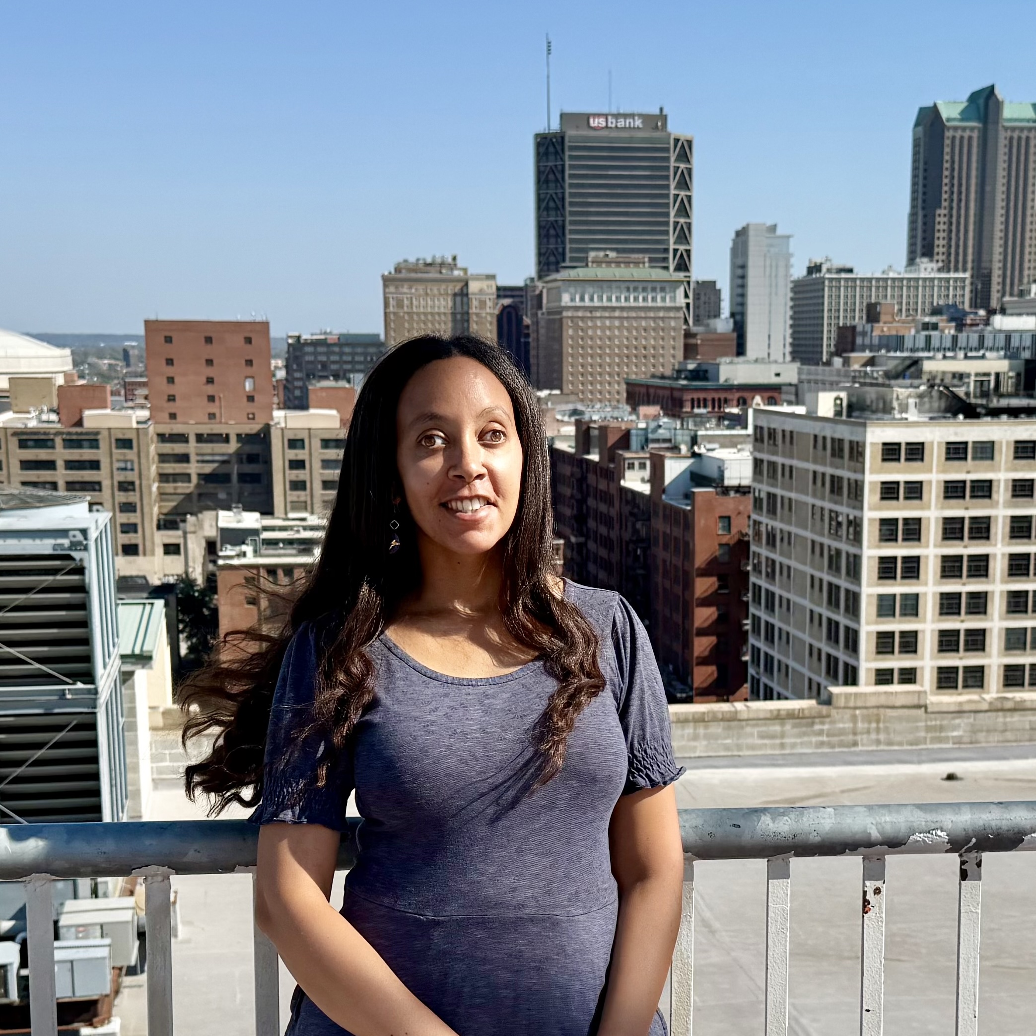 Haben stands on a rooftop on a sunny day with the Saint Louis skyline in the background. A large building with "U.S. Bank" on it is just over her shoulder. She is wearing a blue dress and smiling.