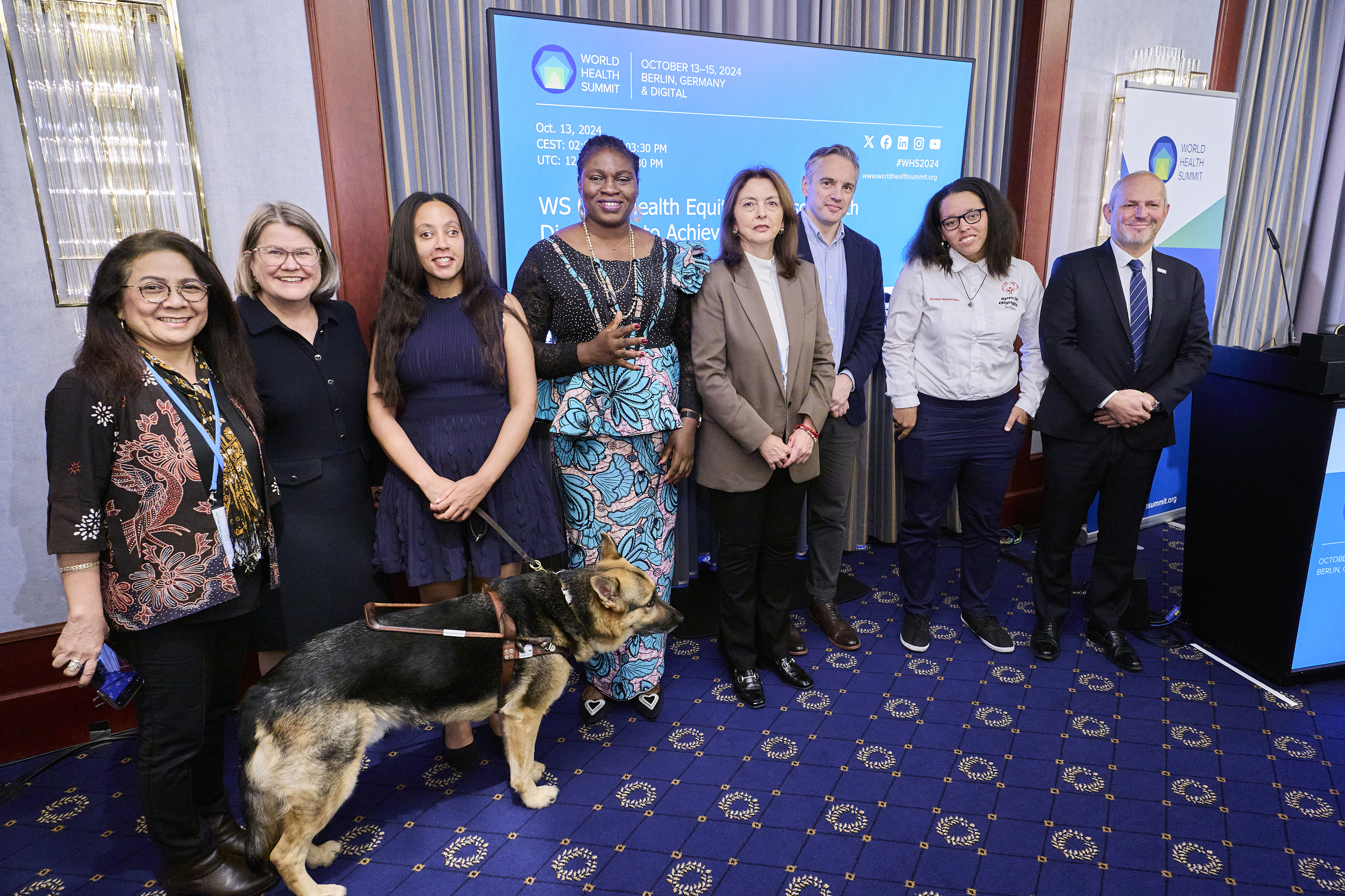 Speakers stand together in front of a blue screen that says “World Health Summit, October 13-15, 2024, Berlin, Germany & Digital.” From left to right we have: R. Vensya Sitohang, Natasha Smith, Haben Girma, Hellen Anurika Beyioku-Alase, María José Plaza, Jarrod Clyne, Rachel Veldkamp, Jérôme Salomon. Haben’s seeing eye dog Mylo is standing in front of her wearing his harness and looking alert, and a podium sits on the far-right side.