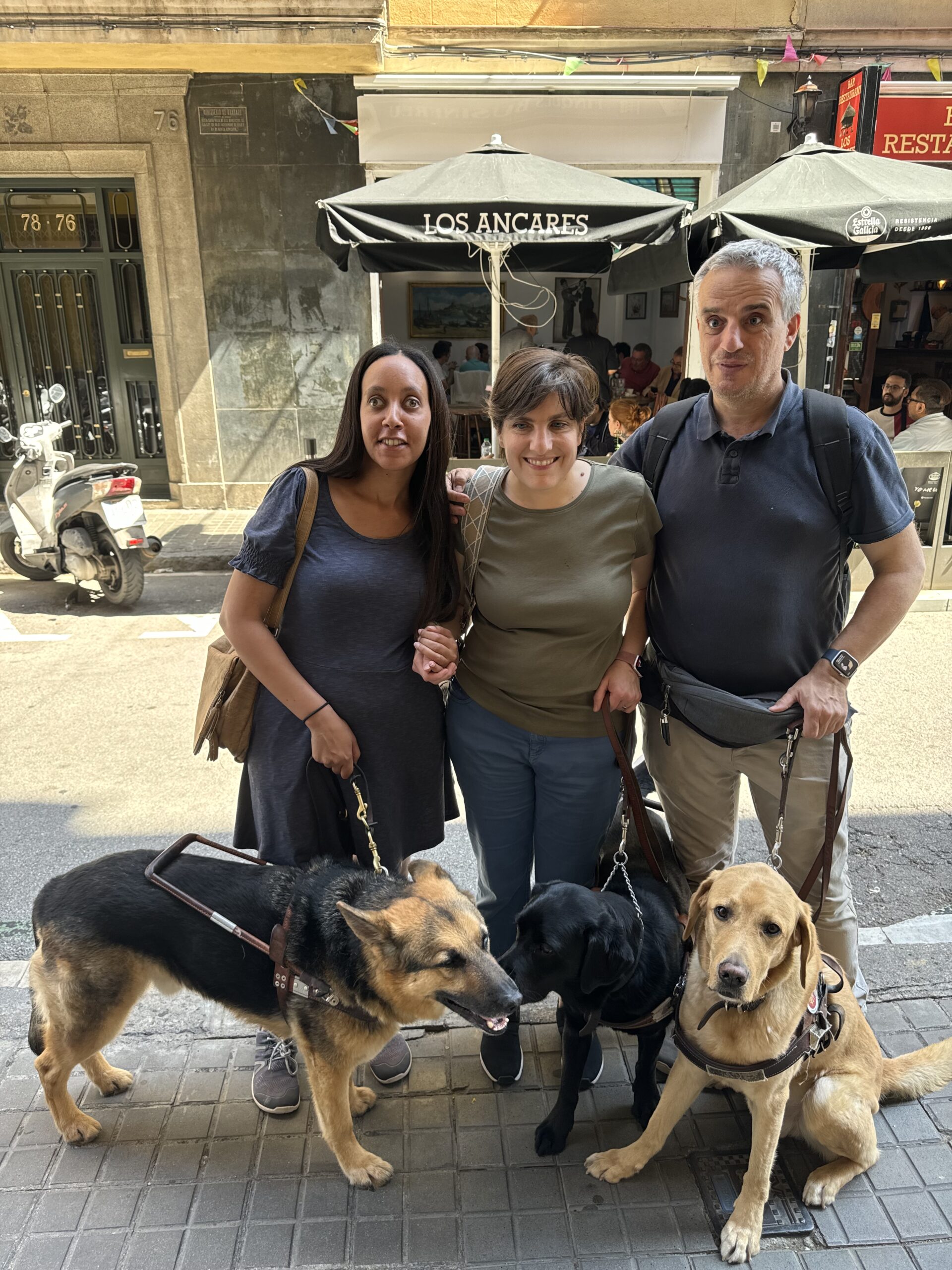 Haben, Daniela, and her husband José stand together on a sunny sidewalk. Haben wears a blue dress, Daniela wears a green shirt and jeans, and José wears a Navy blue shirt, khakis, and an Apple Watch. Next to German Shepherd Mylo is Rosie, a black lab, and beside José sits a yellow lab sweetly looking at the camera. Behind the smiling group, across the street, is a warm restaurant with indoor and outdoor seating.