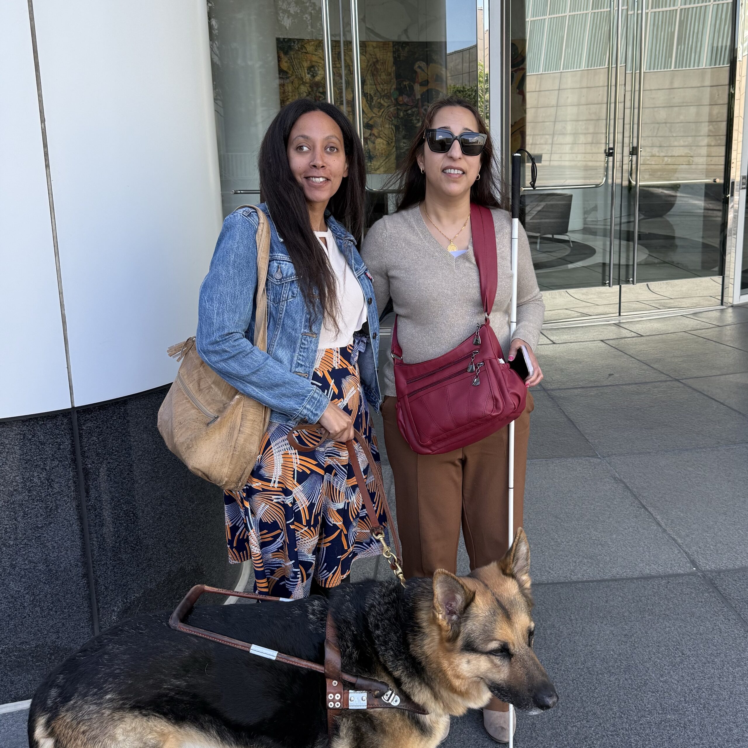 Two women of color stand together smiling. Deepa wears a gold necklace over a gray top, dark yellow trousers, sunglasses, and holds her cane and iPhone in her left hand. Haben wears a denim jacket over a colorful dress and holds the leash of her Seeing Eye dog in her right hand. Behind them is a glass door reflecting the trees in front of the building.