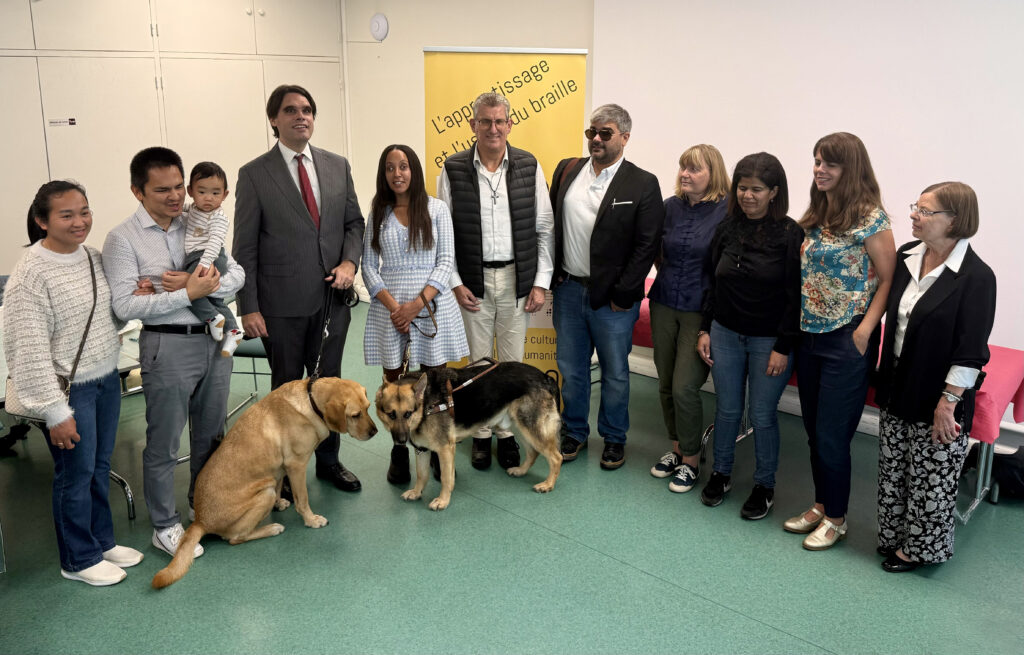A group of people smiling in front of the same sign. From left to right: Pissin Than with his wife and adorable baby, Matthieu Juglar, Rider, Haben Girma, Mylo, Anthony Martins-Misse, Père Hervé Rollin, Sylvie Thézé, Fadila Ouzlifi, Murielle Mauguin, and Christine Hardy.