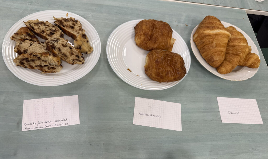 A line of plates full of delicious pastries sit on a table, and each one has a card with Braille and print. From left to right: Brioche with tonka bean and chocolate, Chocolate bread (Pain au chocolat), and croissant.
