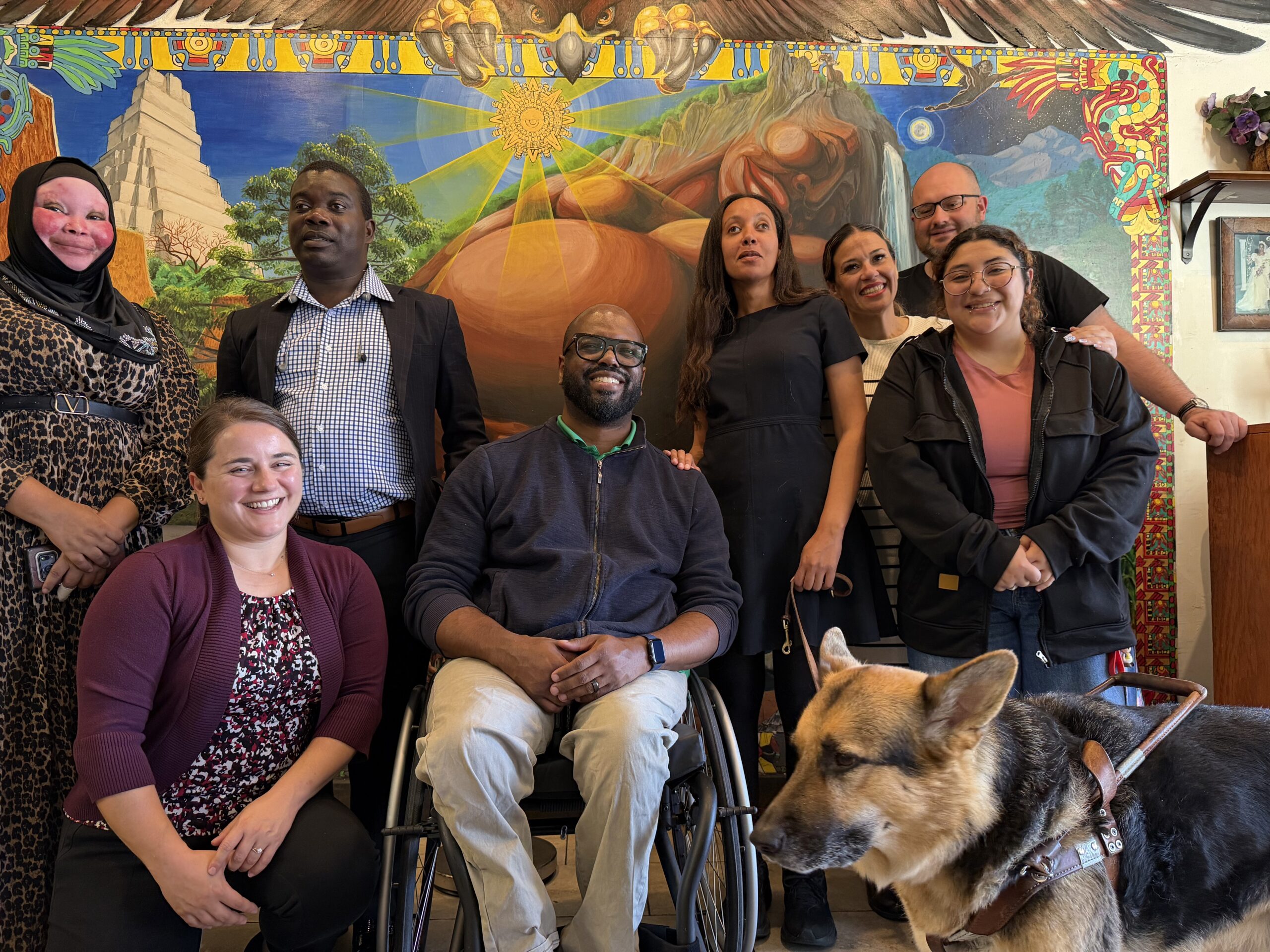 A group of people smiling together in front of a mural with a radiant sun. From left to right we have: Ajjara Ally wearing a leopard print dress and hijab, Rajab Mpilipili in a dark suit jacket over a checkered shirt, Monica Porter Gilbert kneeling beside Eric Harris who sits in a manual wheelchair, Haben Girma has one hand on his shoulder and the other holding her Seeing Eye dog’s leash, Vanessa Ochoa standing with her adult daughter Paris, and Gordon Byrnes wearing a dark shirt and glasses.